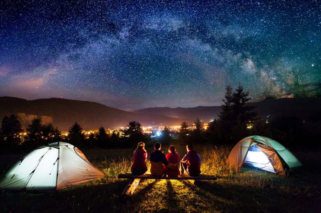 Friends hikers sitting on a bench made of logs and watching fire together beside camp and tents in the night. On the background beautiful starry sky, mountains and luminous town. Rear view