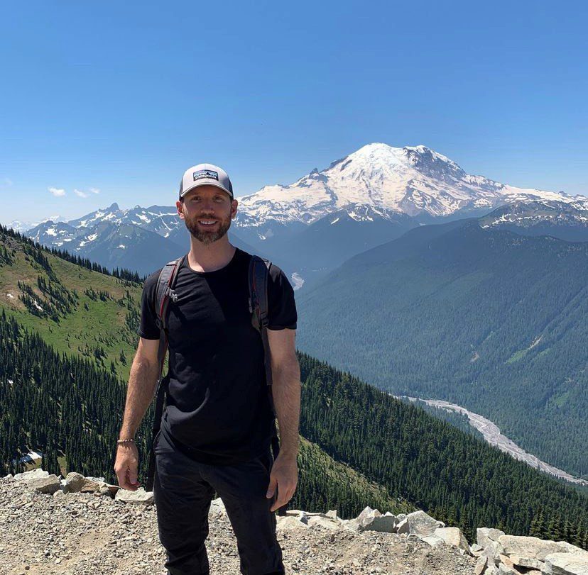 Singing a Travel Nurse’s Praises Man in dark clothing and baseball cap stands in front of mountain landscape