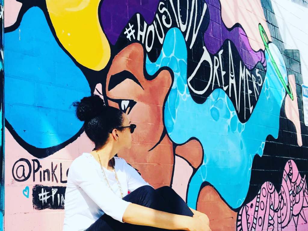 A TNAA employee sitting in front of a vibrant mural located on Leeland Street in Houston, Texas. The mural is created by an artist known as @PinkLomein.