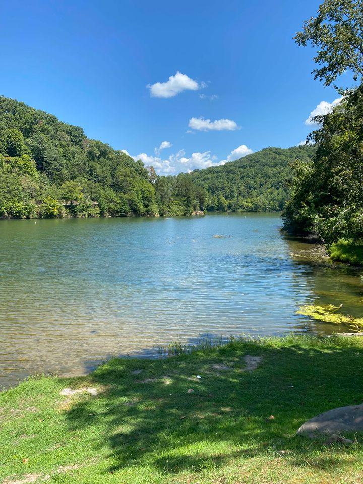 Fishpond Lake near Jenkins, Kentucky. The surrounding area features green, tree-covered hillsides and a clear blue sky with some clouds. The webpage text is a blog post titled "Must-See Places in Kentucky on Your Travel Assignment", which discusses various natural attractions in the state, including Mammoth Cave National Park, Cumberland Falls State Resort Park, and Red River Gorge. 